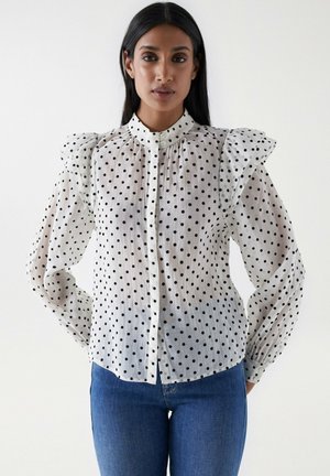 Blusa blanca con lunares, de cuello alto, con hombros acampanados y cierre de botones. Tejido transparente con mangas largas abullonadas. Combinada con jeans azules.