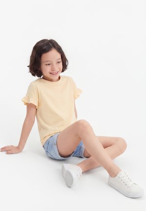 Fată tânără stând pe podea, purtând un top galben cu mâneci scurte, pantaloni scurți deschiși la culoare din denim și teniși albi, zâmbind și uitându-se în jos.