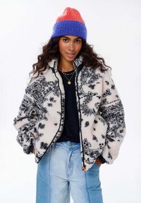 Fleece bunda v krémovej farbe s čiernym kvetinovým vzorom, so zipsom a bočnými vreckami; spárovaná s multikolórovou pletenou čiapkou.