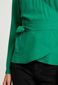 Grüne Wickelbluse mit langen Ärmeln, Bindegürtel in der Taille und einer fließenden Silhouette. Der Stoff erscheint glatt, leicht und strukturiert.
