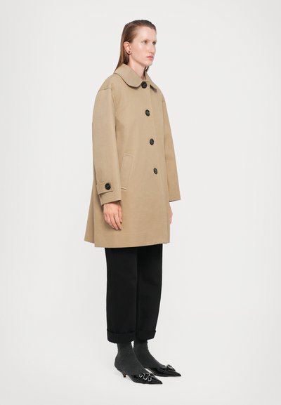 Cappotto color sabbia, lungo fino al ginocchio, con un ampio colletto, bottoni neri e tasche laterali, abbinato a pantaloni neri con risvolto e scarpe con punta.