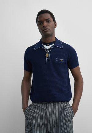 Man with hands in pockets wearing navy short-sleeve knit polo, striped pants, and layered gold medallion necklaces against gray background.