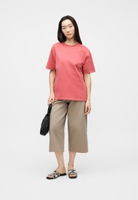 Γυναίκα που στέκεται φορώντας ένα ξεθωριασμένο κόκκινο μπλουζάκι, φαρδιά μπεζ παντελόνια μέχρι τον αστράγαλο, σανδάλια με μοτίβο φιδιού και κρατώντας μια μικρή μαύρη τσάντα χειρός.