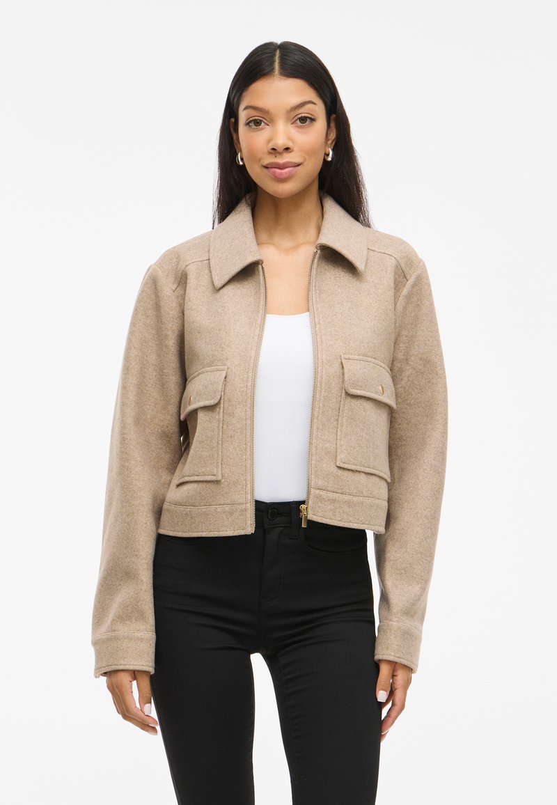 Giacca corta beige in morbida lana, con colletto, due tasche frontali e chiusura a zip. Abbinata a una maglietta bianca e pantaloni neri.