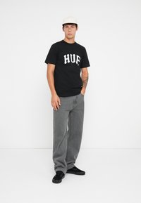 Fekete pamut póló fehér "HUF" felirattal, szürke, bő szabású farmernadrággal és fekete sportcipővel, sima fehér háttér előtt.