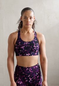 Juodas sportinis liemenukas su violetiniais gėlių raštais ir kryžminiu nugaros dizainu, derinamas su aukštu juosmeniu atitinkančiais leggings. Lygus, elastingas audinys.