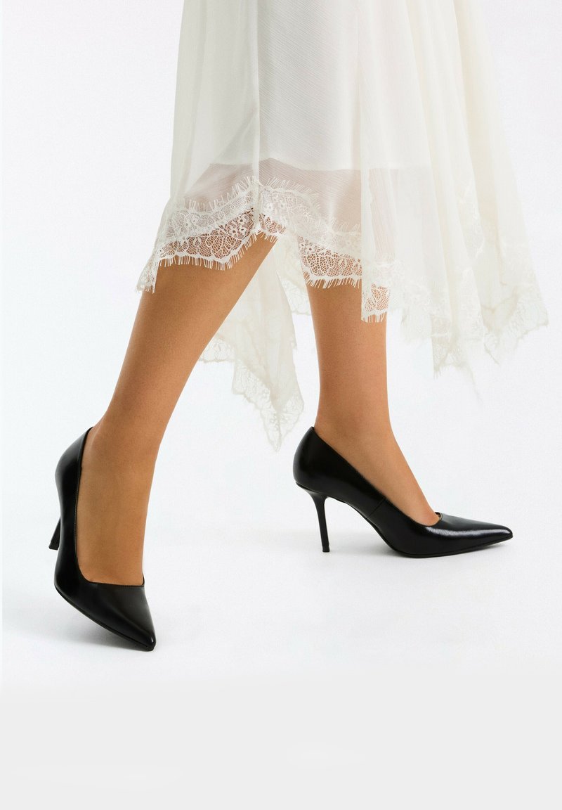 Jambes portant des bas beige transparents et des escarpins noirs à bout pointu sous une robe blanche bordée de dentelle.