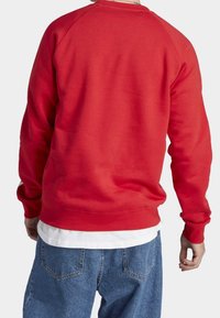 Felpa rossa in cotone con maniche raglan, tinta unita, polsini e orlo a coste, indossata sopra una T-shirt bianca, abbinata a jeans denim blu.