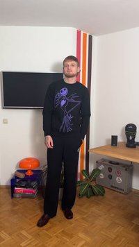 Μαύρο φούτερ με γραφικό σκίτσο μωβ σκελετού, συνδυασμένο με μαύρο παντελόνι και σκούρα παπούτσια. Το περιβάλλον περιλαμβάνει πολύχρωμες ριγέ τοίχους και διακοσμητικά στοιχεία.