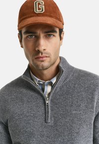Grijze ziptrui met geribde kraag, gelaagd over een blauw-wit gestreept shirt. Bruine corduroy pet met een gouden letter "G."
