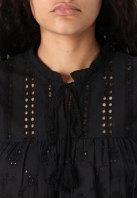 Blusa nera con colletto arricciato, legature frontali e dettagli in pizzo. Il tessuto è leggero, con orlo arricciato e accenti testurizzati.