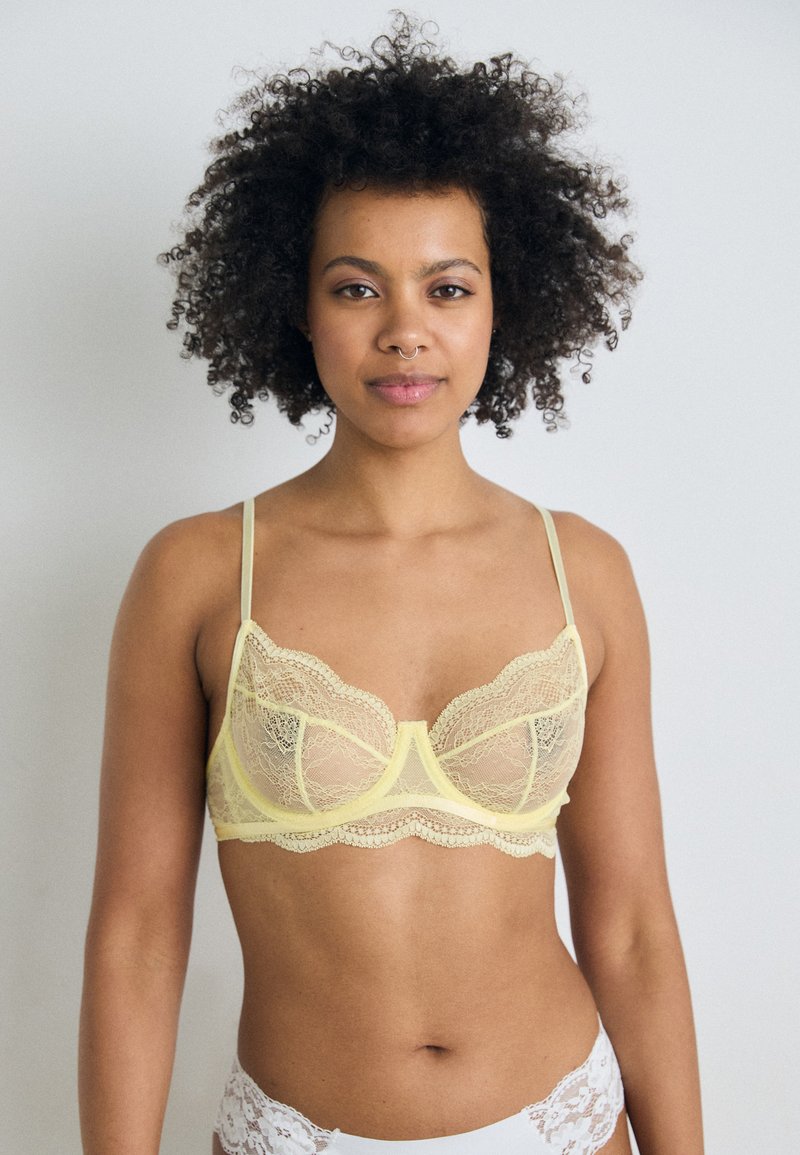 Lace bra in pale yellow with scalloped edges, adjustable shoulder straps, and underwire support, featuring a delicate, sheer texture.