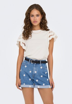 Jeune femme aux cheveux bouclés portant un haut blanc à manches volantes, un short en jean imprimé de marguerites blanches, une ceinture noire et des boucles d'oreilles en or en forme de créole.