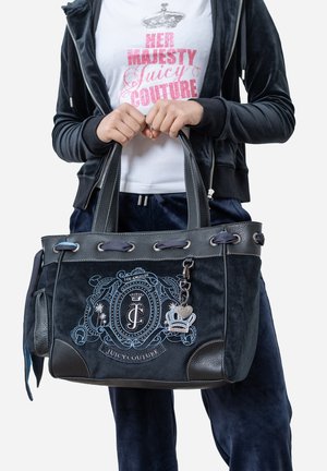 Handen die een zwarte Juicy Couture tas vasthouden met een blauw geborduurd schild, gecombineerd met een donker fluwelen outfit en een wit T-shirt met "Her Majesty Juicy Couture".