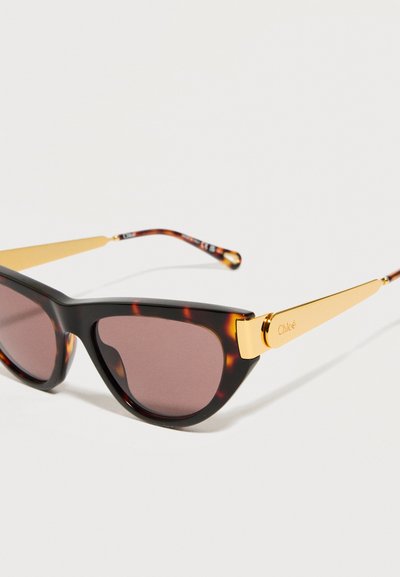 Γυαλιά ηλίου τύπου cat-eye με σχέδιο από χελώνα, σκουρόχρωμους φακούς και χρυσά πλαϊνά, με το όνομα της μάρκας χαραγμένο στον βραχίονα.