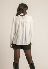 Blouse blanche à manches longues avec un dos plissé, associée à un short noir, des collants transparents et des bottes noires jusqu'aux genoux.