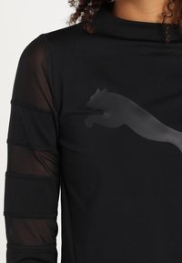 Musta pitkähihaisuus paita, jossa on läpinäkyvät verkko- ja vaakaraidat. Etupuolella on matta musta Puma-logo. Sujuva kankaan pinta.