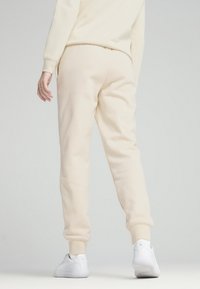 Beige joggerbroeken gemaakt van zacht, textuurmateriaal met elastische boorden. Gecombineerd met witte sneakers, gezien vanuit de achterkant.