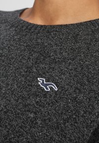 Grå stickad tröja med ett strukturerat mönster, med en liten vit- och marinhavsblå logotyp på vänster bröstområde. Rund halsringning.