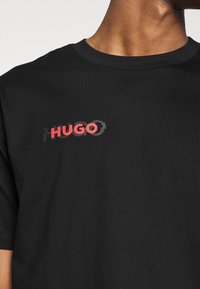 Černé bavlněné tričko s výrazným grafickým logem "HUGO" v červené a šedé barvě. Kulatý výstřih a krátké rukávy s měkkou texturou.