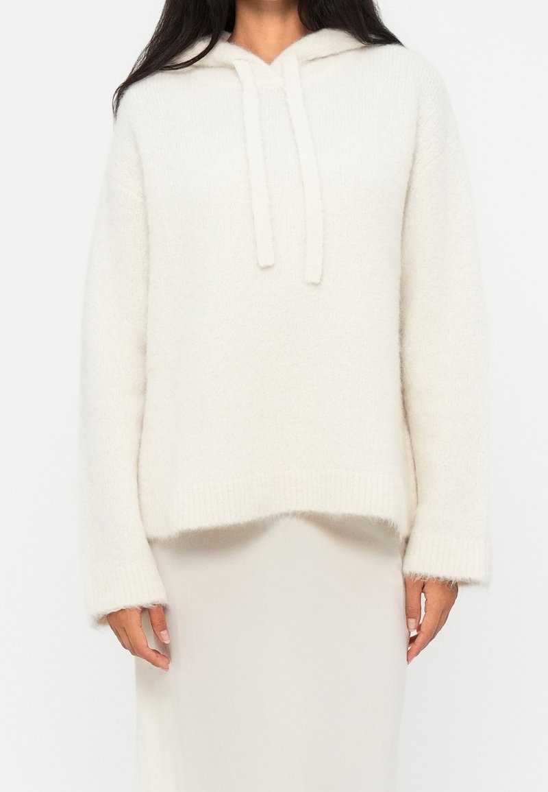 Femme portant un pull à capuche blanc doux à manches longues, associé à une jupe de couleur claire, debout devant un fond uni.