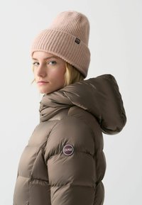 Bruine pufferjas met capuchon en gestikte ontwerpen, met een cirkelvormig Colmar-logo op de linkermouw. Roze geribde beanie op het model.