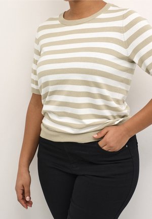 Persoon draagt een beige-wit gestreept shirt met korte mouwen en een zwarte broek, met één hand in de zak en de andere ontspannen langs het lichaam.