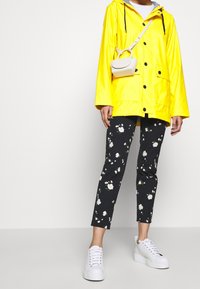Żółta wodoodporna kurtka z czarnymi guzikami i kapturem, połączona z czarnymi spodniami w kwiatowy wzór oraz białymi tenisówkami. Mała torebka na ramię.