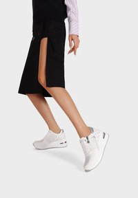 Sneakers atletici bianchi con tomaia texture, dettagli metallici argentati e suola ammortizzata. Indossati con una gonna nera dotata di uno spacco laterale.