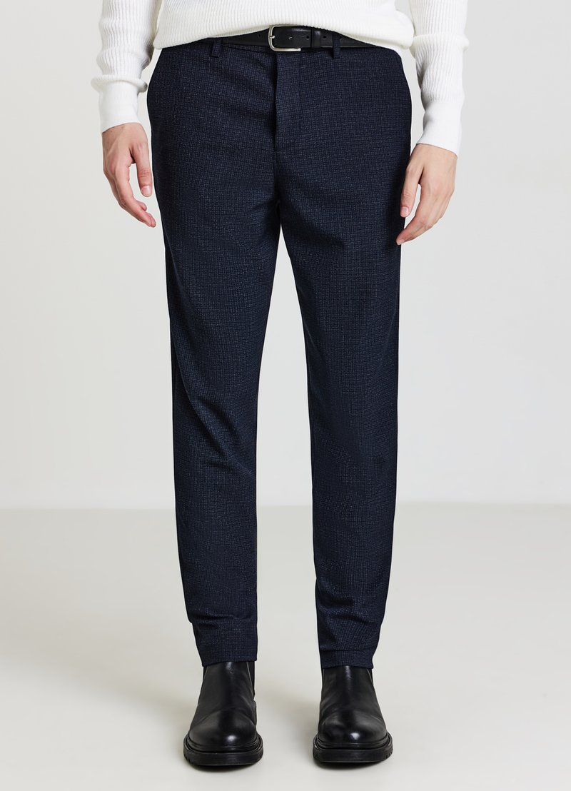 Dunkelblaue, gemusterte Hose mit schmaler Passform aus strukturiertem Stoff, kombiniert mit schwarzen Leder-Stiefeletten und einem weißen Strickpullover.