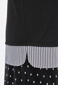 T shirt nera con orlo arrotondato indossata sopra una camicia a righe grigie e bianche, con una texture lucida e una gonna nera con pois bianchi.