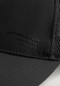 Schwarze Mütze aus strukturiertem Stoff mit Netzseiten und geprägtem Detail "UNIFORM UTILITY EXPO 0073" an der Vorderseite.