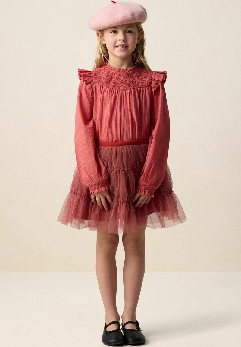 Robe rouge avec manches à volants et jupe en tulle à volants. Associée à un béret rose en feutre et des chaussures noires. Détails de tissus lisses et texturés.