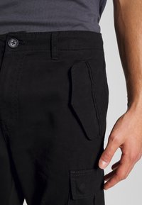 Pantalons cargo noirs en tissu résistant, dotés d'une poche latérale et de coutures visibles. Gros plan sur la taille et la main sur la poche.