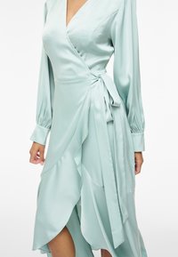 Φόρεμα τύπου wrap από σατέν ανοιχτού γαλάζιου χρώματος με μακριά φουσκωτά μανίκια, V-λαιμόκοψη και δεμένη φιόγκο στη μέση, παρουσιασμένο σε μοντέλο από τον λαιμό έως τη μέση της γάμπας.