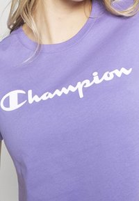 Lila bomull t-shirt med en vit skriplogotyp "Champion" över bröstet. Korta ärmar och rundad halsringning.