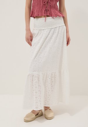 Donna che indossa una gonna lunga maxi in pizzo bianco a occhielli con vita arricciata, espadrillas beige e top rosso corto senza maniche con laccetti.