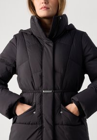 Γυναίκα που φοράει μια μαύρη επλυσμένη χειμερινή καμπαρντίνα με ψηλό γιακά και τα χέρια της στις κουμπωμένες μπροστινές τσέπες, με φόντο απλό.