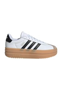 Zapatilla deportiva blanca con un talón negro texturizado, tres rayas negras y una suela de plataforma marrón. Presenta materiales de cuero liso y tela.