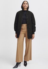 Jachetă bomber neagră cu manșete ribbate, asociată cu pantaloni largi bej și pantofi cu vârf ascuțit negri. Textură netedă, design ajustat și închidere cu fermoar.