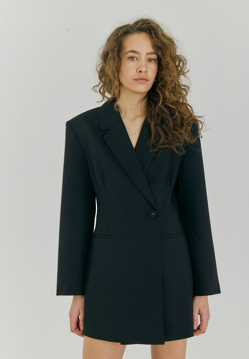 Čierne oversized blazer šaty s jedným gombíkom, ostrými chlopňami, dlhými rukávmi a upravenými líniami. Hladká tkanina so štruktúrovaným strihom.