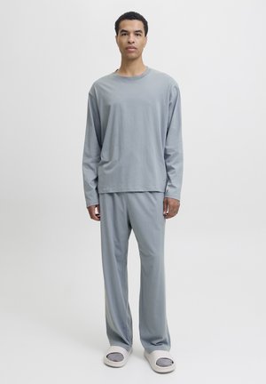 Mann in einem lockeren, grauen Langarmshirt und grauen Hosen mit grauen Socken und weißen Badesandalen, der vor einem schlichten weißen Hintergrund steht.