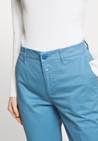 Ljusblå bomullsbukser med flat front, dragkedja och rund knappstängning. Har sidofickor och en liten logotyp.