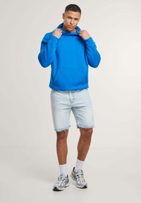 Felpa blu brillante con tasca frontale, abbinata a pantaloni corti in denim chiaro. Il modello indossa calzini bianchi a crew e sneakers grigie con dettagli blu.