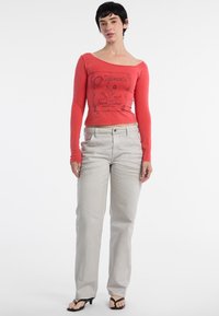 Rotes Langarmoberteil mit grafischem Druck; hellgraue gerade Hose; das Model trägt schwarze Flip-Flops; einfaches, figurnahes Design.
