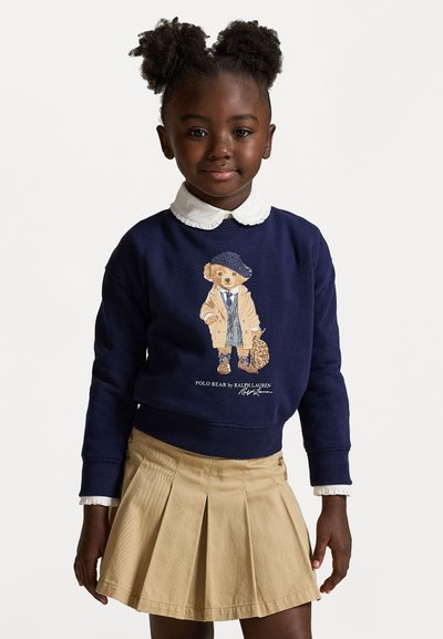 Navy sweatshirt med en björn grafik i en trench coat, kombinerad med en ljusbeige plisserad kjol. Vit skjorta med krage under.