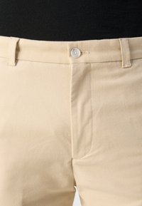 Pantalon beige avec fermeture à boutons et passants pour ceinture, porté avec un haut noir, montrant la taille et la partie supérieure des cuisses de face.