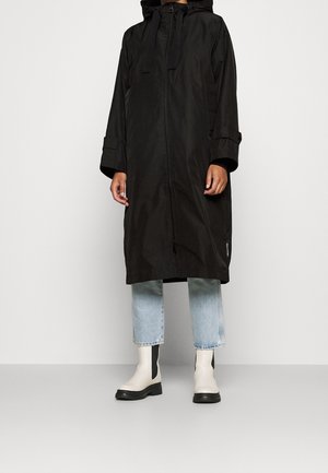 Personne portant un long manteau noir à capuche avec une fermeture éclair à l'avant, un jean court bleu clair et des bottines blanches avec des semelles noires.