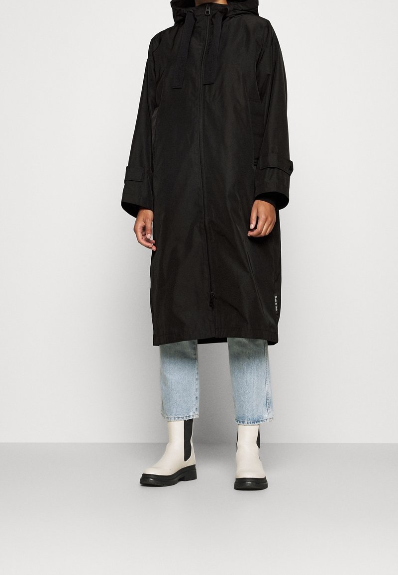 Personne portant un long manteau noir à capuche avec une fermeture éclair à l'avant, un jean court bleu clair et des bottines blanches avec des semelles noires.