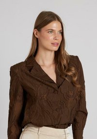 Blusa marrone con motivo, dal finish testurizzato, caratterizzata da un colletto a punte e chiusura a bottoni. Tessuto morbido con dettagli di design sottotono.